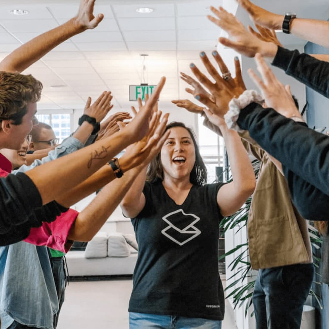 Enthusiastic group celebrating with raised hands in an office space, woman in black shirt with logo at centre.